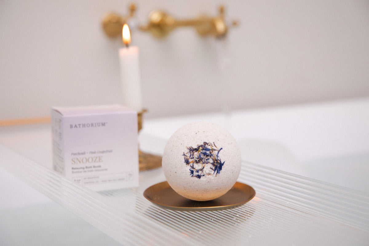 Bathorium Snooze Bath Bomb topped with dried petals on a brass dish beside its box and a candle.