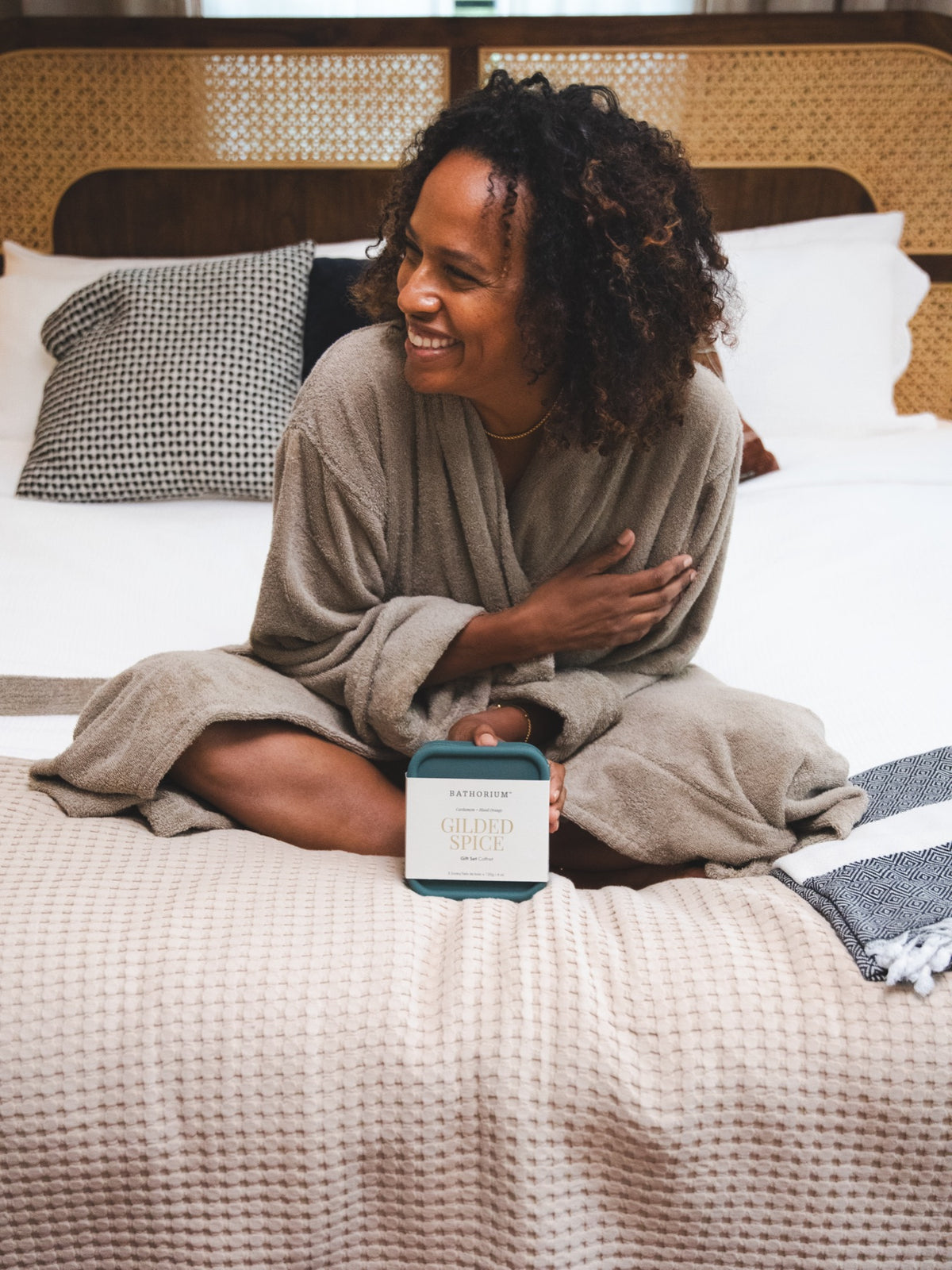 Laughing on the bed in a robe, holding the Gilded Spice Travel Kit